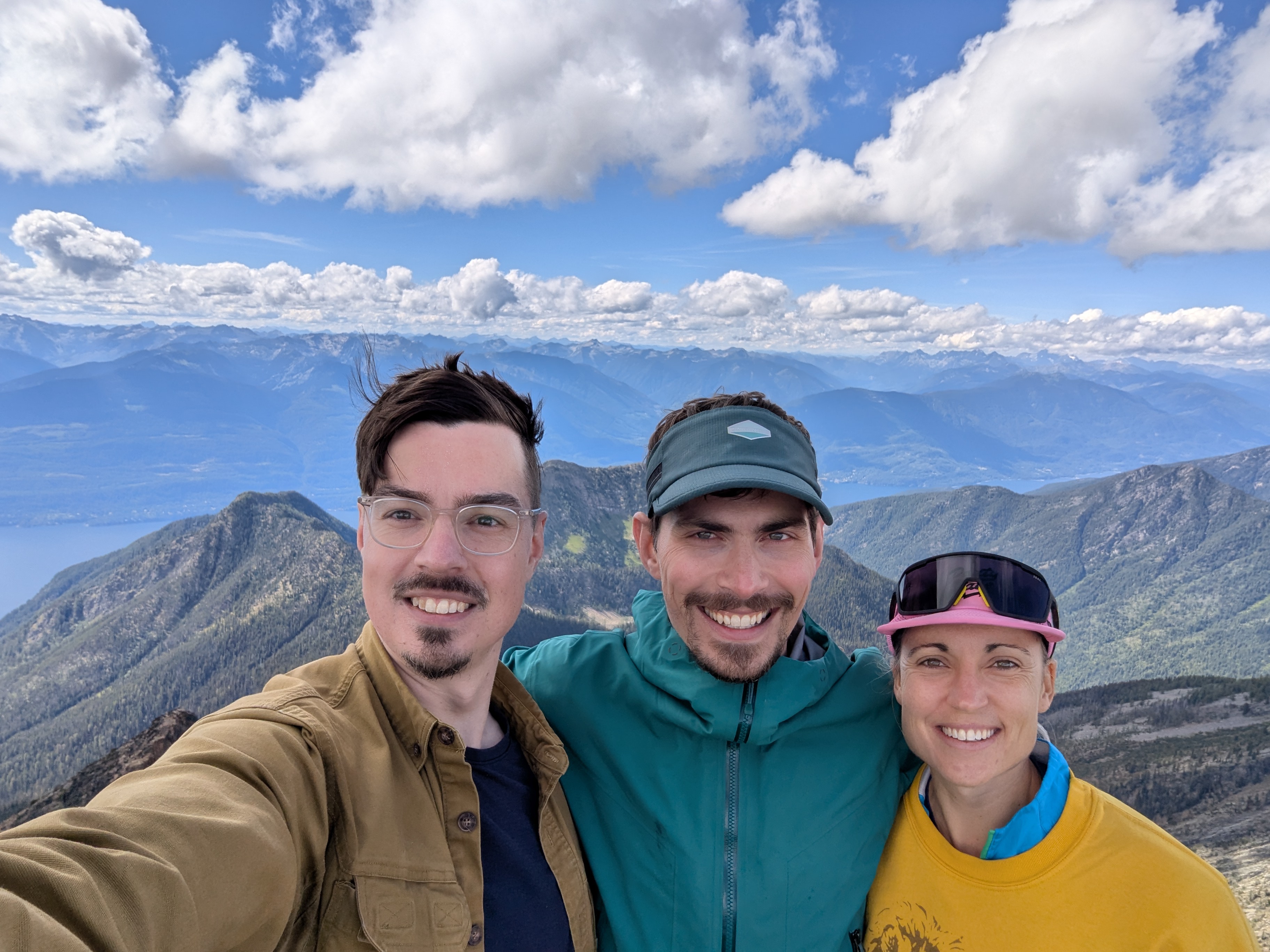 Three people at the top of a mountain. The image includes a blue partly cloudy sky with a backdrop of mountainous territory.